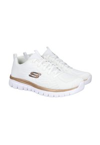 Par de zapatillas deportivas blancas ligeras con suelas acolchadas, diseño con cordones y detalle en bronce en el lateral y la entresuela.