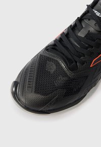 Črne atletske čevlje z drsno površino, črnimi vezalkami in oranžnimi detajli na belem ozadju, nagnjeni, da pokažejo podrobnosti prstov in zgornjega dela.