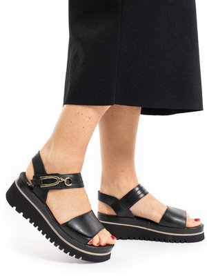 Piedi con smalto rosso sulle unghie dei piedi che indossano sandali neri con plateau, cinturini alla caviglia e fibbia dorata, abbinati a pantaloni neri.