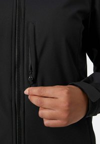 Zwarte softshell jas met een ritssluiting op de borstzak, met een gladde textuur, een slim fit ontwerp en verstelbare manchetten. Hand grijpt de rits van de zak.