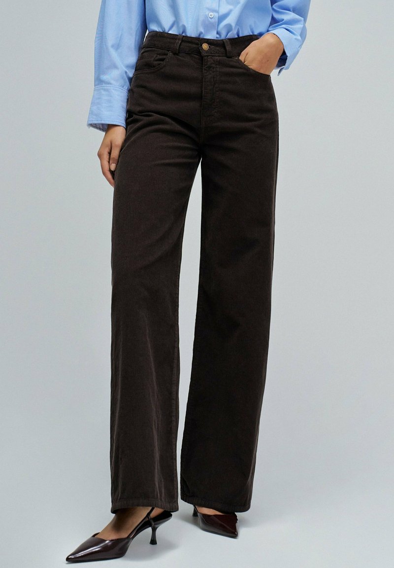 Pantalones anchos de pana marrón con cintura alta, que cuentan con bolsillos frontales y cierre de botón. Combinados con tacones negros de punta.