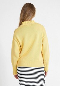 Gelber Pullover mit strukturiertem Strick, Volantkragen und gerippten Bündchen. Über einem gestreiften schwarz-weißen Rock getragen.