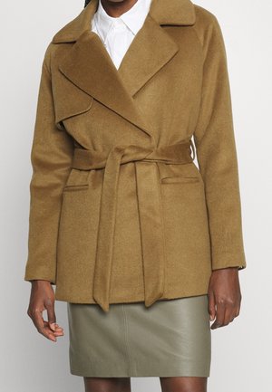 Kobieta w płaszczu z wełny w kolorze beżowym z paskiem, na białej koszuli z kołnierzykiem i oliwkowo-zielonej skórzanej spódnicy, stojąca na jednolitym tle.