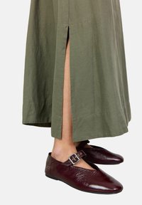 Scarpe in pelle borgogna con punta affilata, cinturino regolabile con fibbia, indossate con un vestito verde oliva fino alla caviglia con fessure laterali.