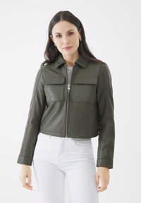 Abgeschnittene olivgrüne Lederjacke mit zwei Fronttaschen, einem Reißverschluss und einem Kragen. Tragen über einem hellfarbenen Oberteil und weißen Hosen.