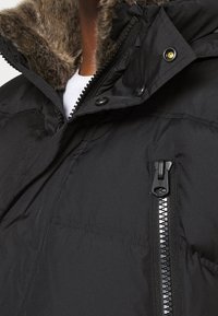 Sort puffjakke med en pelsforet hætte, lynlåslukning foran og markante syninger. Materialet ser glat og tekstureret ud.