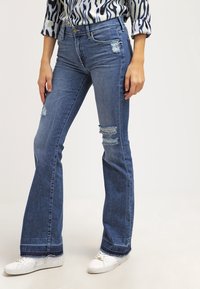 Rozšířené džíny v středně modrém denimu s odřenými detaily, s vysokým pasem a třepícími se lemy. Vamilé s bílými teniskami.