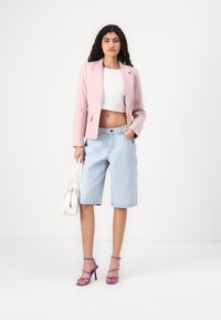 Světle růžový blazer přes bílý crop top, doplněný světle modrými denimovými kraťasy a fialovými sandály na podpatku. Drží bílou kabelku.