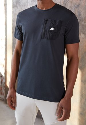 Mann trägt schwarzes Nike-T-Shirt mit Reißverschlusstasche auf der Brust und weißen Hosen, steht drinnen vor einer strukturierten Wand.