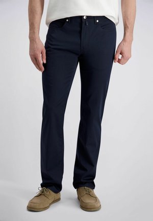 Homme portant un pantalon droit bleu marine foncé et des chaussures beiges en daim à lacets, debout devant un fond gris clair.