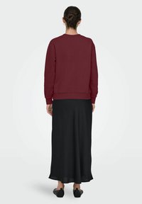 Vrouw met een bun, gekleed in een bordeauxrode sweatshirt, een zwarte lange rok en zwarte platte schoenen, staande met haar rug naar de camera op een effen achtergrond.