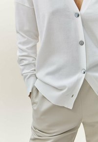 White cardigan made of soft fabric, featuring a v-neck, four grey buttons, and ribbed cuffs, paired with beige trousers.