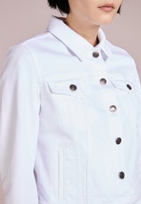 Osoba ubrana w białą kurtkę dżinsową z metalowymi guzikami i klapowymi kieszeniami na piersi, widoczna od ramion do połowy tułowia na neutralnym tle.
