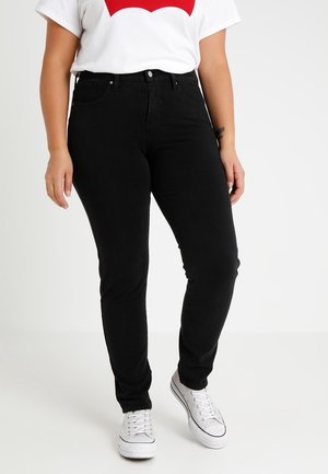 Person trägt schwarze Slim-Fit-Jeans, weiße Freizeitsneaker und ein weißes T-Shirt mit rotem Motiv vor schlichtem Hintergrund.