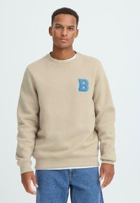 Beige mikina s kulatým výstřihem, vyrobená z měkkého materiálu; má na hrudi modro vyšívané logo "B" a manžety se žebrováním.