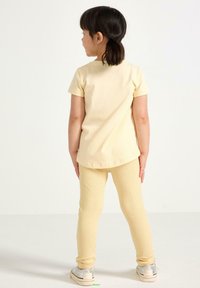 Gul bomulds T-shirt med korte ærmer og rund kant, parret med ribbede, lys gule leggings. Modellen har sølvfarvede sko på.
