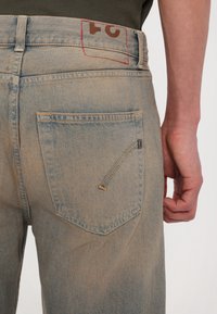 Gros plan d'une personne portant un jean en denim bleu clair délavé montrant la poche arrière et la ceinture avec un motif cousu rouge.
