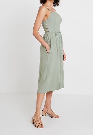 Nainen, joka käyttää hihattomaa salvianvihreää midimekkoa, jossa on sivulla napit ja taskut, yhdistettynä beigeihin avojalkaisiin lohkopiikkikorollisiin sandaaleihin.