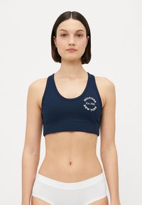 Marineblå sports-BH med rund halsutskjæring, med en hvit logo som sier "HILFIGER EST. 1985 NEW YORK," laget av et glatt stoff.