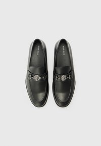 Mocasines de cuero negro con una textura suave, que cuentan con una hebilla de acento plateado y detalle de costura en la punta. Nombre de la marca visible en el interior.