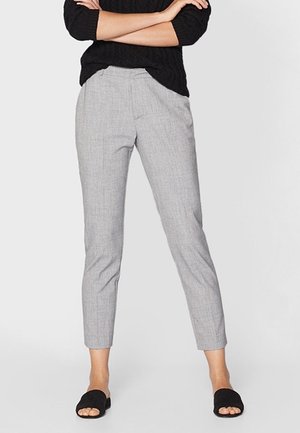 Persoon draagt lichtgrijze op maat gemaakte enkellange broek, zwarte gebreide trui, zwarte platte sandalen met open tenen, staat met gekruiste armen.