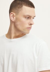 White cotton T-shirt with a crew neckline, featuring a subtle embroidered logo on the chest. Smooth texture, short sleeves.