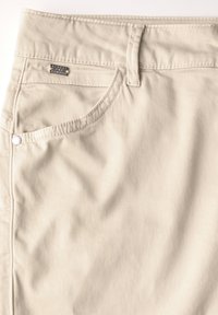 Pantalons en coton beige avec une texture lisse, une poche avant et des éléments en métal avec une étiquette de marque à l'arrière.