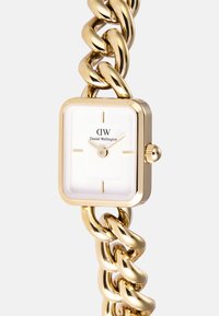 Montre dorée avec un cadran blanc carré, arborant un logo de marque noir, des index horaires minimalistes et un bracelet en chaîne dorée épais.