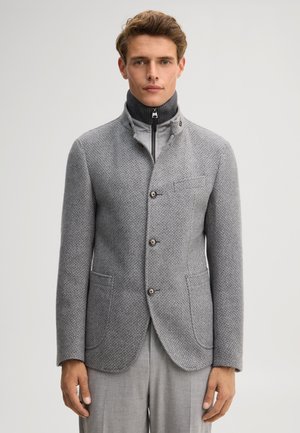 Man draagt een gestructureerde grijze knoopblazer over een grijze coltrui met rits en bijpassende grijze broek, staand tegen een effen achtergrond.