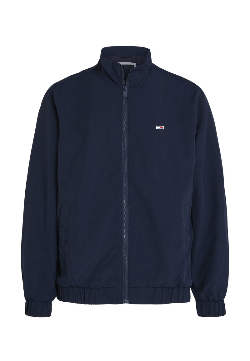Veste légère bleu marine avec un col montant, des poignets et un ourlet élastiques, et un petit logo sur la poitrine gauche. Fermeture à glissière sur toute la longueur des devants.
