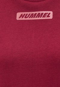 Gros plan sur un tissu bordeaux avec le logo rose "HUMMEL" imprimé près de l'encolure sur un vêtement tricoté.
