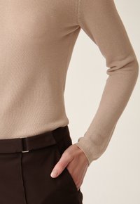 Haut à manches longues en tricot beige clair avec un tissu texturé, associé à un pantalon tailleur marron foncé avec un accent métallique subtil à la taille.
