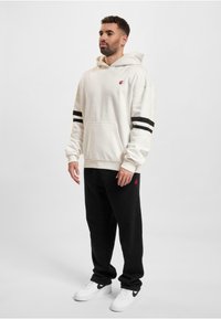 Felpa con cappuccio color crema con strisce nere sulle maniche, dotata di tasca frontale e logo rosso. Abbinata a pantaloni della tuta neri e sneakers bianche.