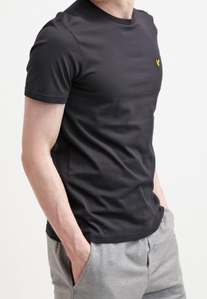 Homme portant un t-shirt noir uni à manches courtes avec un petit logo jaune sur la poitrine, et un pantalon de survêtement gris clair, les mains dans les poches.
