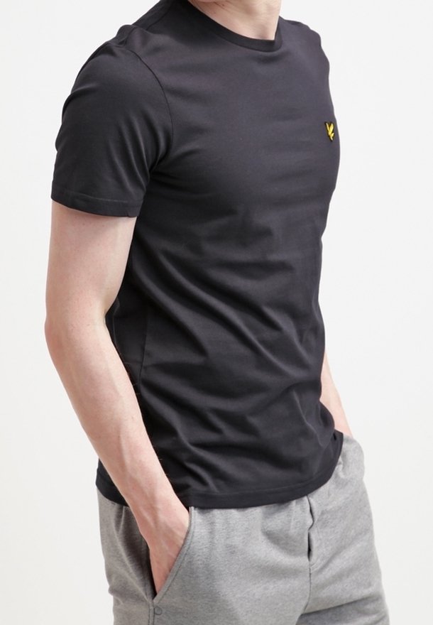 Homme portant un t-shirt noir uni à manches courtes avec un petit logo jaune sur la poitrine, et un pantalon de survêtement gris clair, les mains dans les poches.
