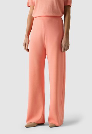 Kvinde iført koralfarvede bukser med høj talje og vide ben og en matchende kortærmet top, stående foran en ensartet lys baggrund.