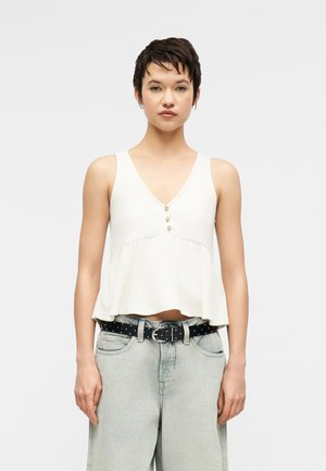 Νεαρός με κοντά σκούρα μαλλιά φοράει αμάνικο λευκό πουκάμισο με κουμπιά και ανοιχτόχρωμο τζιν με μαύρη ζώνη με τρουκς, στέκεται μπροστά σε απλό φόντο.