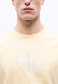 Jeune adulte portant un t-shirt Calvin Klein jaune pâle avec un grand logo "cK" et un texte centré sur la poitrine, fond blanc.