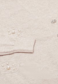Lichtbeige cardigan gemaakt van zacht breisel. Heeft parelaccenten en een decoratieve knop op de voorkant en de manchetten.