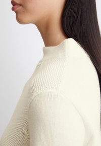 Maglione di lana crema con collo alto, design strutturato sulle spalle e tessuto del corpo liscio. Il colore è morbido e neutro.