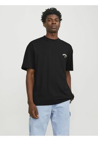 Sort bomulds T-shirt med rund hals og korte ærmer, prydet med et lille hvidt grafisk design på venstre brystområde. Båret med lyseblå jeans.