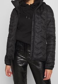 Giubbotto imbottito nero con silhouette aderente, chiusura con zip e cappuccio. Tessuto testurizzato con motivo trapuntato e polsini elasticizzati.