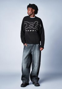 Czarny dziergany sweter z białym tekstem graficznym i falistym wzorem, zestawiony z luźnymi, ciemnymi jeansami. Model nosi czarne trampki.