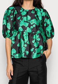 Donna che indossa una blusa nera con maniche a sbuffo e motivi floreali verde brillante, abbinata a pantaloni neri, con una mano in tasca.