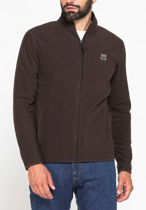 Uomo che indossa una giacca in pile marrone scuro con cerniera e colletto alto, e jeans blu, mostrato dalle spalle ai fianchi.