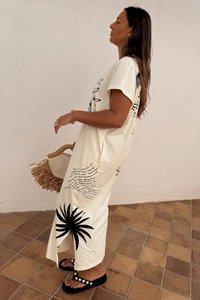 Abito t-shirt oversize color bianco sporco con testo nero e stampa di palme. Presenta spacchi ai lati, abbinato a sandali neri e una borsa di paglia.