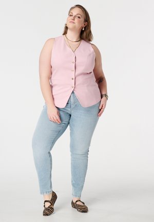 Zelfverzekerde vrouw staat met een mouwloos roze knoopvestje, lichtblauwe jeans en platte schoenen met tijgerstrepen tegen een effen achtergrond.