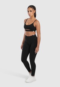 Brasile sportivo nero con dettagli a taglio e spalline sottili, abbinato a leggings neri a vita alta. Tessuto liscio, design aderente, indossato con sneakers bianche.