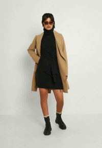 Cardigã longo bege, vestido preto de gola alta com cinto, e botas de combate pretas. A modelo usa óculos de sol oversized e meias caneladas em creme.