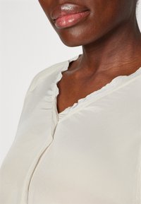 Blouse de couleur crème avec une texture douce, présentant un col en dentelle et des coutures détaillées le long des bords.